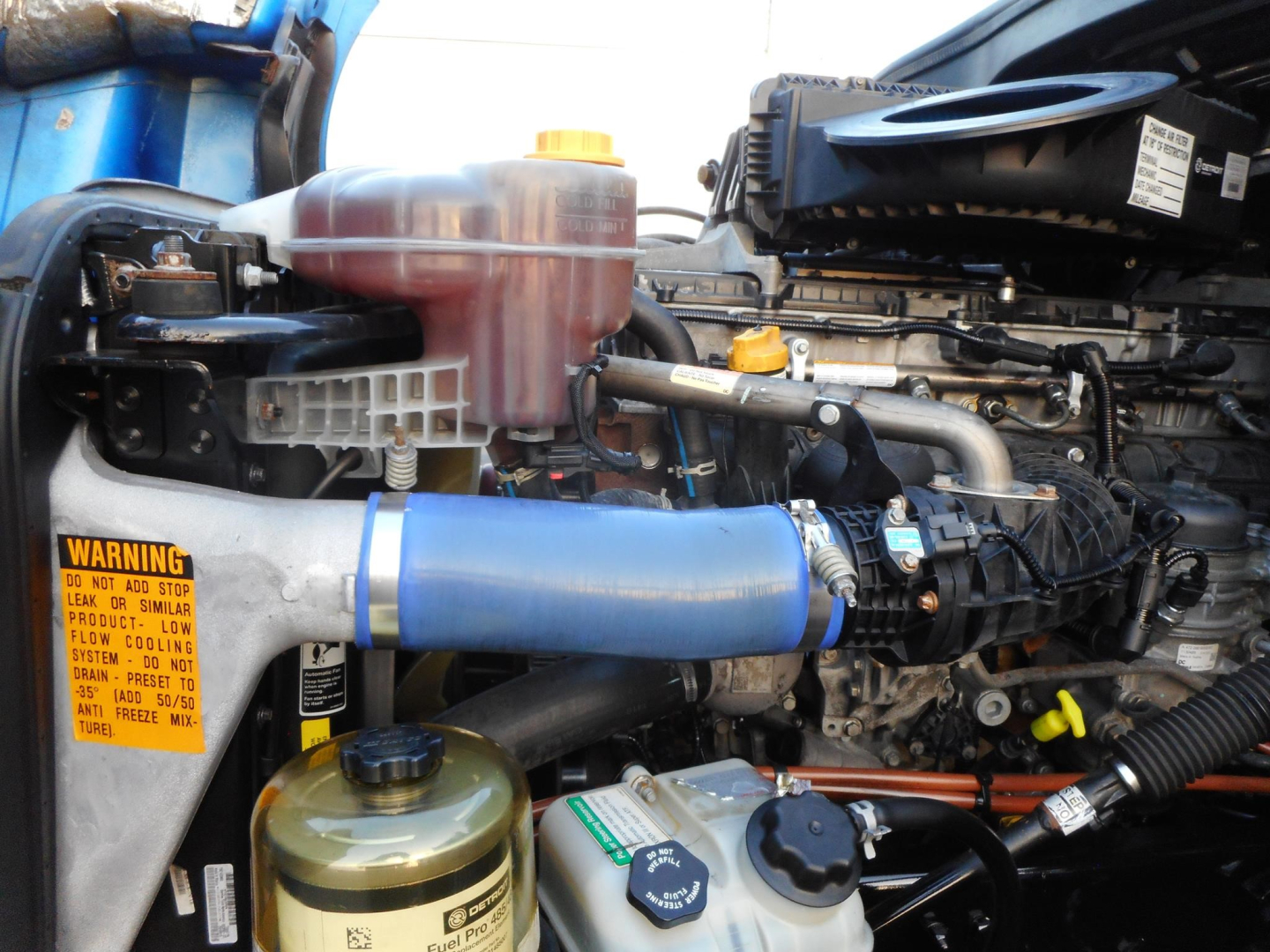 Engine bay of Freightliner Cascadia showing coolant reservoir, pink expansion tank, blue intercooler hose, and various hoses and clamps.
