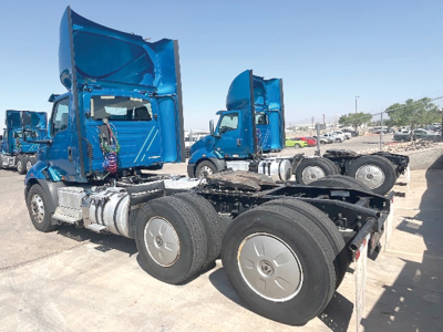 Truck chassis and cabs of a 2019 International RH613 conventional daycab with blue paint, dual rear axles, exposed fuel tanks, and multiple similar trucks in background.