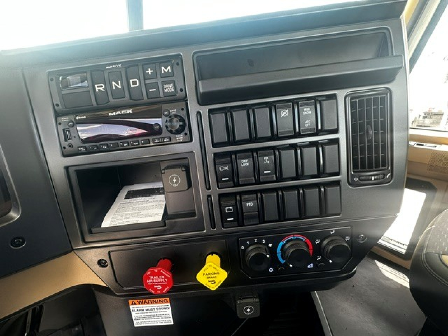 Interior of a Mack GR64F dash showing gear lever area (R N D P) and multiple switch panels, gauges, and climate controls in the dashboard.