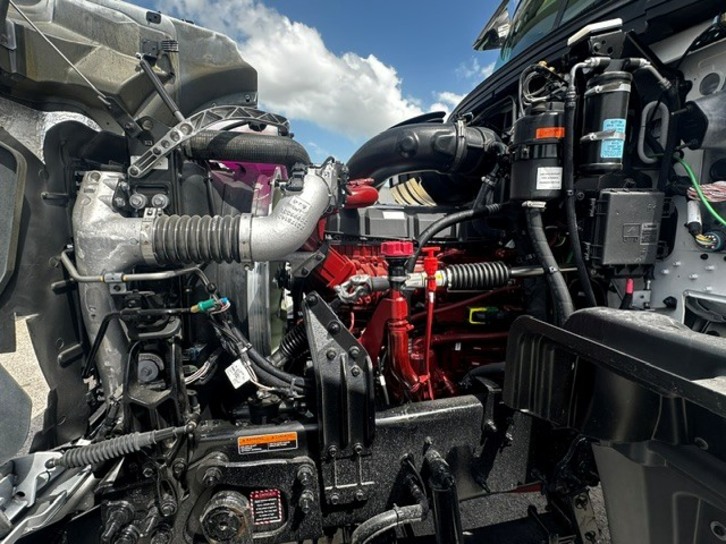 Truck engine bay showing Mack GR64F components with red engine parts and various hoses and electricals.