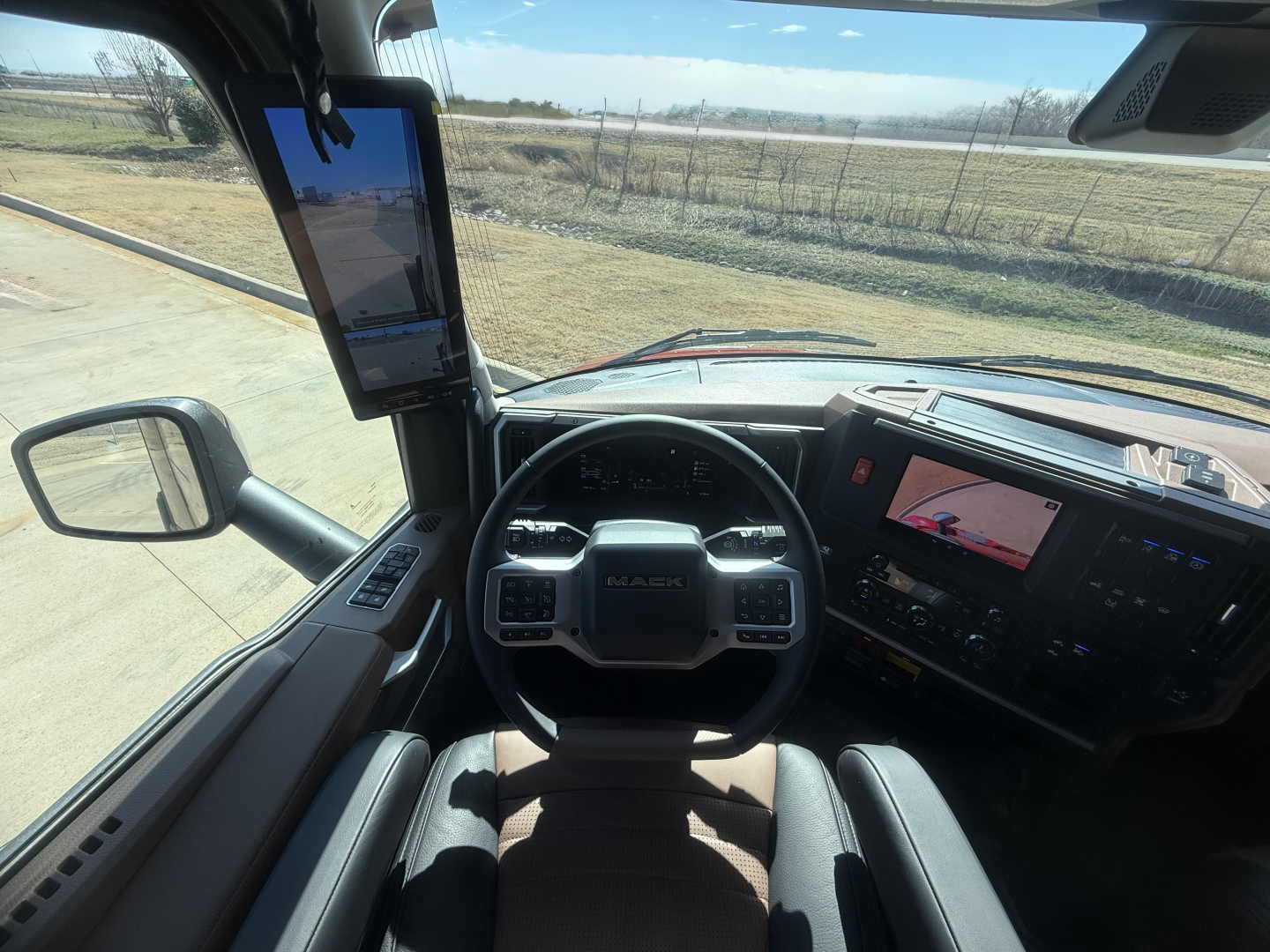 Truck cockpit view of a 2026 Mack PR64T76H with steering wheel, dashboard, and large side mirror. The driver area shows a tablet mounted on the A-pillar and a center touchscreen display.