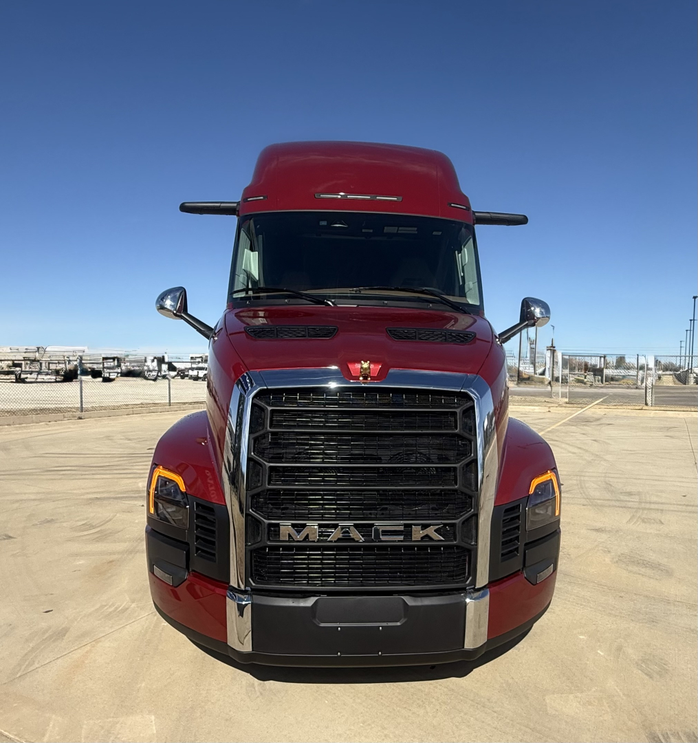 Front view of a red 2026 Mack PR64T76H truck showing bold grille, headlights, chrome bumper, Mack emblem, and wide windshield in a parking lot.