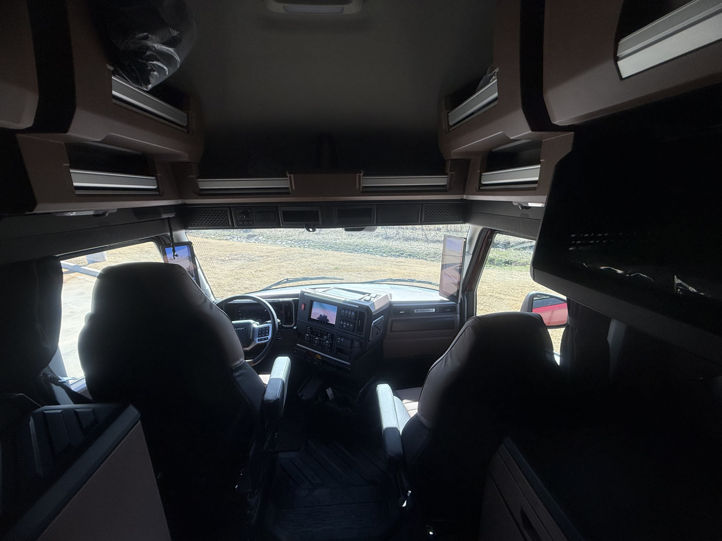 Interior cab view of a 2026 Mack PR64T76H showing overhead sleeper storage cabinets and dashboard with two front seats.