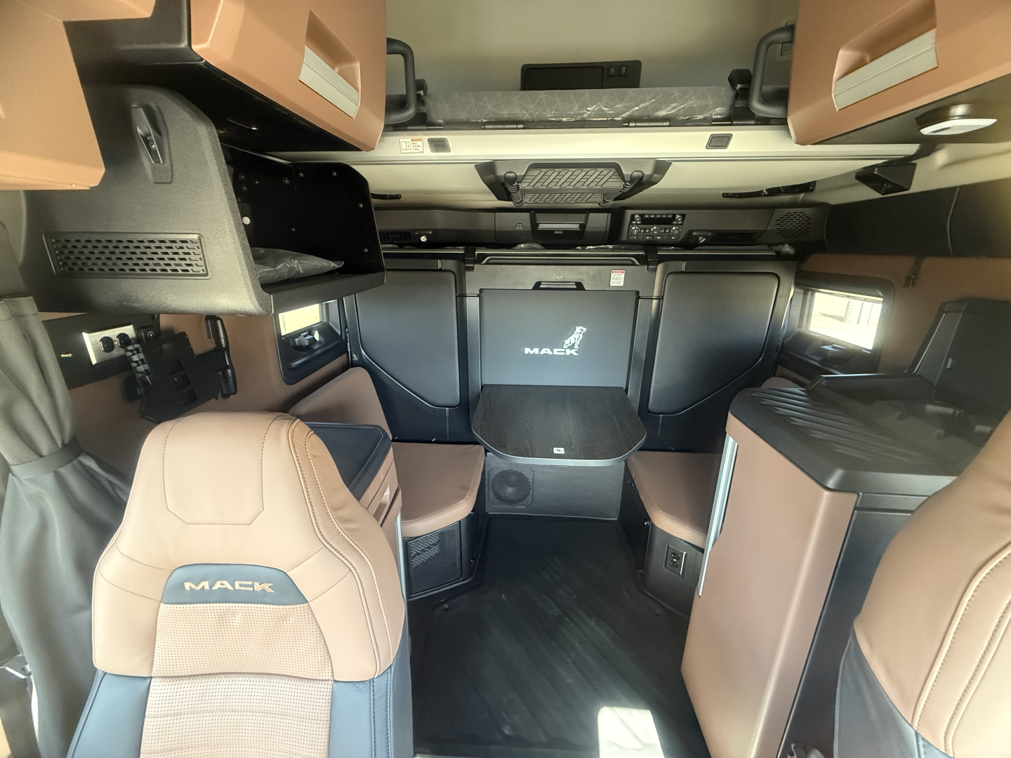 Interior of a new Mack PR64T76H sleeper cab showing tan and black leather seats, overhead storage, and dashboard area with Mack logo on partition.