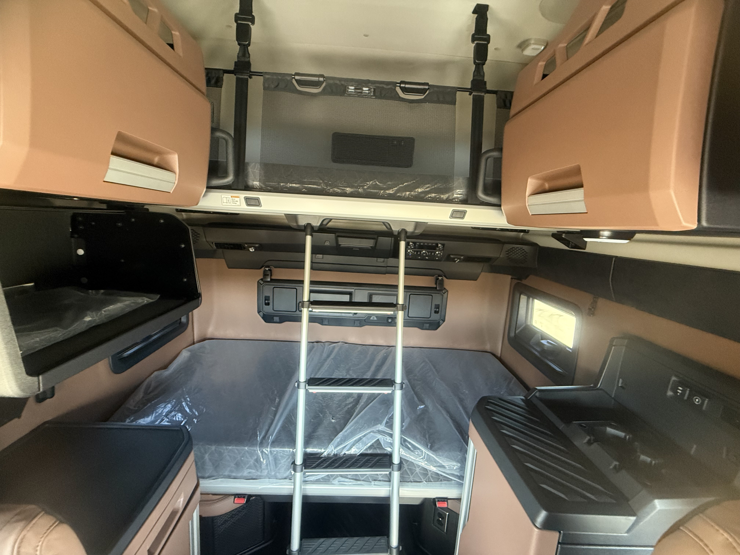 Cab interior view of a 2026 Mack PR64T76H sleeper cab showing upper storage pods, ladder to bunk area, and dashboard/pass-through with beige/tan trim.