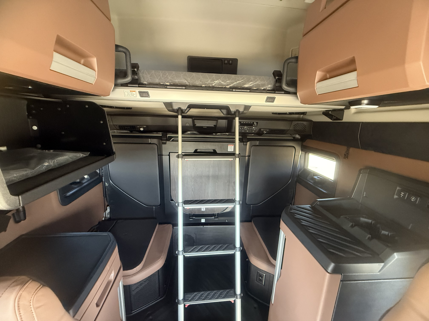 Truck cab interior of a 2026 Mack PR64T76H sleeper showing bunk area, ladder, overhead storage, and side cabinets with tan upholstery.