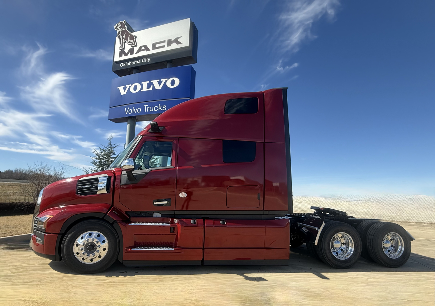 Mack PR64T76H conventional sleeper truck side view showing red exterior, long hood, cab with aero mirrors, tandem rear axles, Volvo Trucks sign in background.