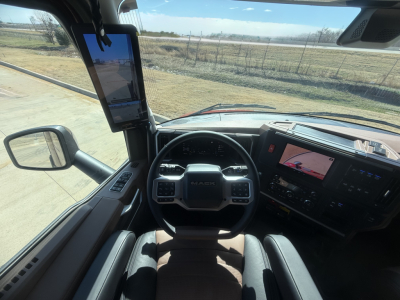 Truck cockpit view of a 2026 Mack PR64T76H with steering wheel, dashboard, and large side mirror. The driver area shows a tablet mounted on the A-pillar and a center touchscreen display.