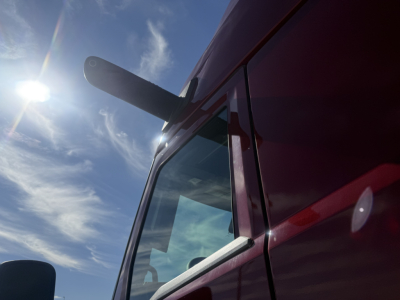 Front cab of a red Mack PR64T76H tractor showing side window, door, and large exhaust stack under bright sun.