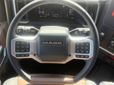Mack PR64T76H truck interior showing steering wheel with Mack emblem and control buttons. Dashboard behind with digital display.