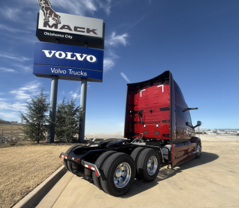 Mack sign and Volvo Trucks sign in front of a red Mack PR64T76H truck, with a clear blue sky. Visible text includes MACK, OKLAHOMA CITY, VOLVO, Volvo Trucks, and a red sleeper cab rear and dual rear axles.