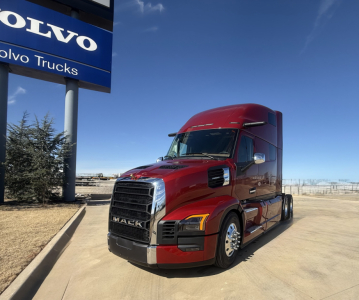 Red 2026 Mack PR64T76H conventional sleeper truck parked at a dealership lot with Volvo sign in background.