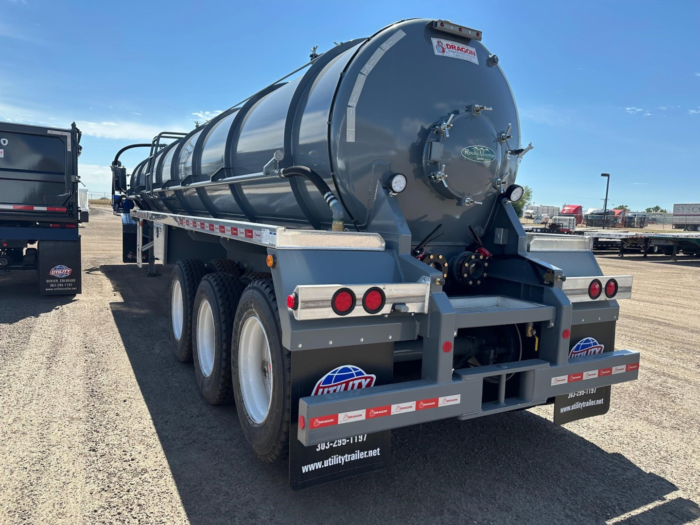 Tank trailer with cylindrical vacuum tank mounted on tri-axle chassis. Rear shows lights, mudguards, and warning labels. Tank has round manhole and hoses.