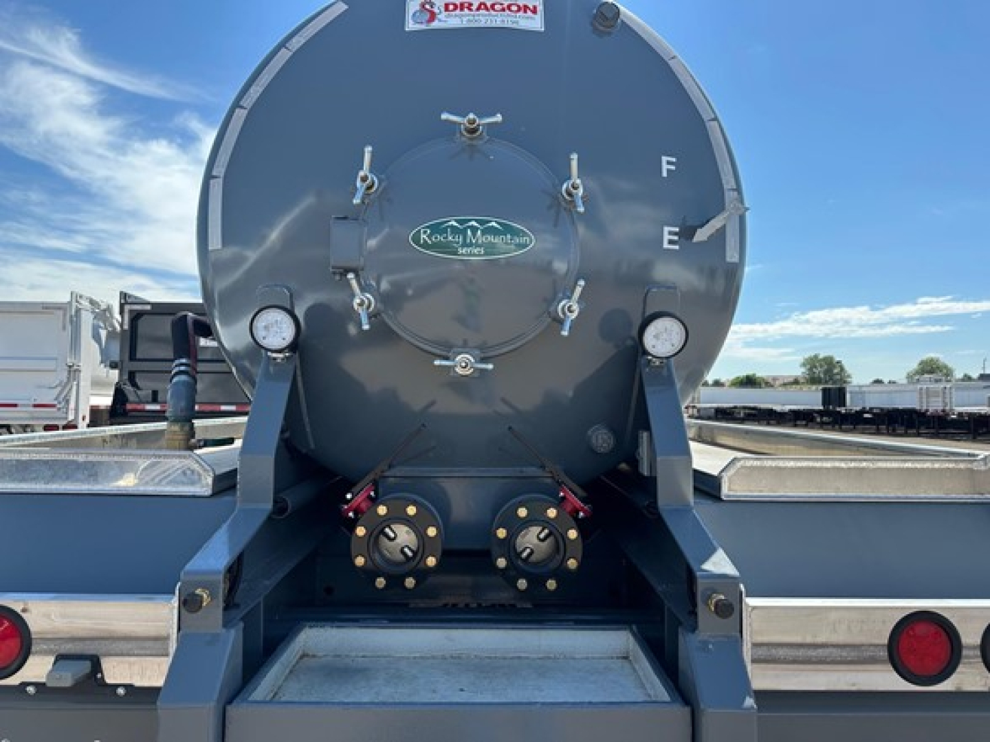 Tank rear view showing circular vacuum tank with doors, gauges, and fittings; brand sticker in center, two bottom hoses, side rails, and rear lights.
