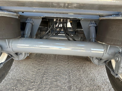 Trailer undercarriage showing a gray hydraulic/dump-style rear axle with dual wheels and a solid central beam; exposed hoses and fittings