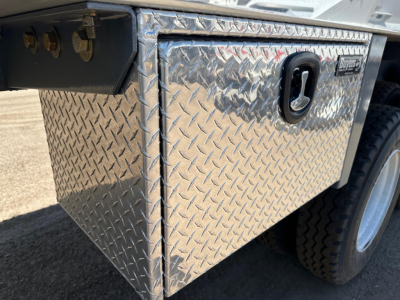 Front left side of a diamond-plate aluminum toolbox mounted on a trailer frame with a latch handle and dual tires visible.