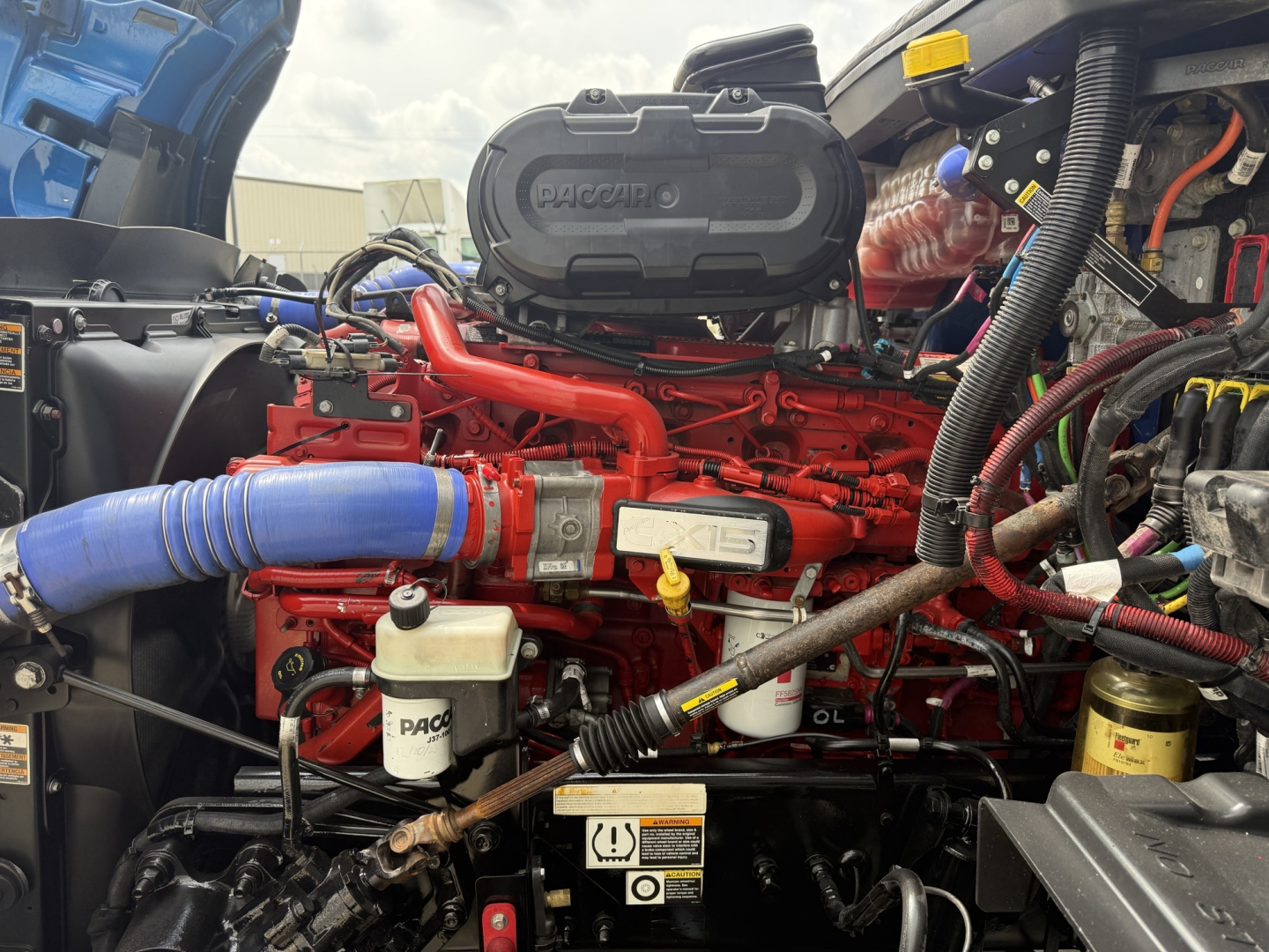 Peterbilt engine bay showing red engine with air intake and blue hose, various hoses, reservoirs, and wiring in a truck engine compartment.