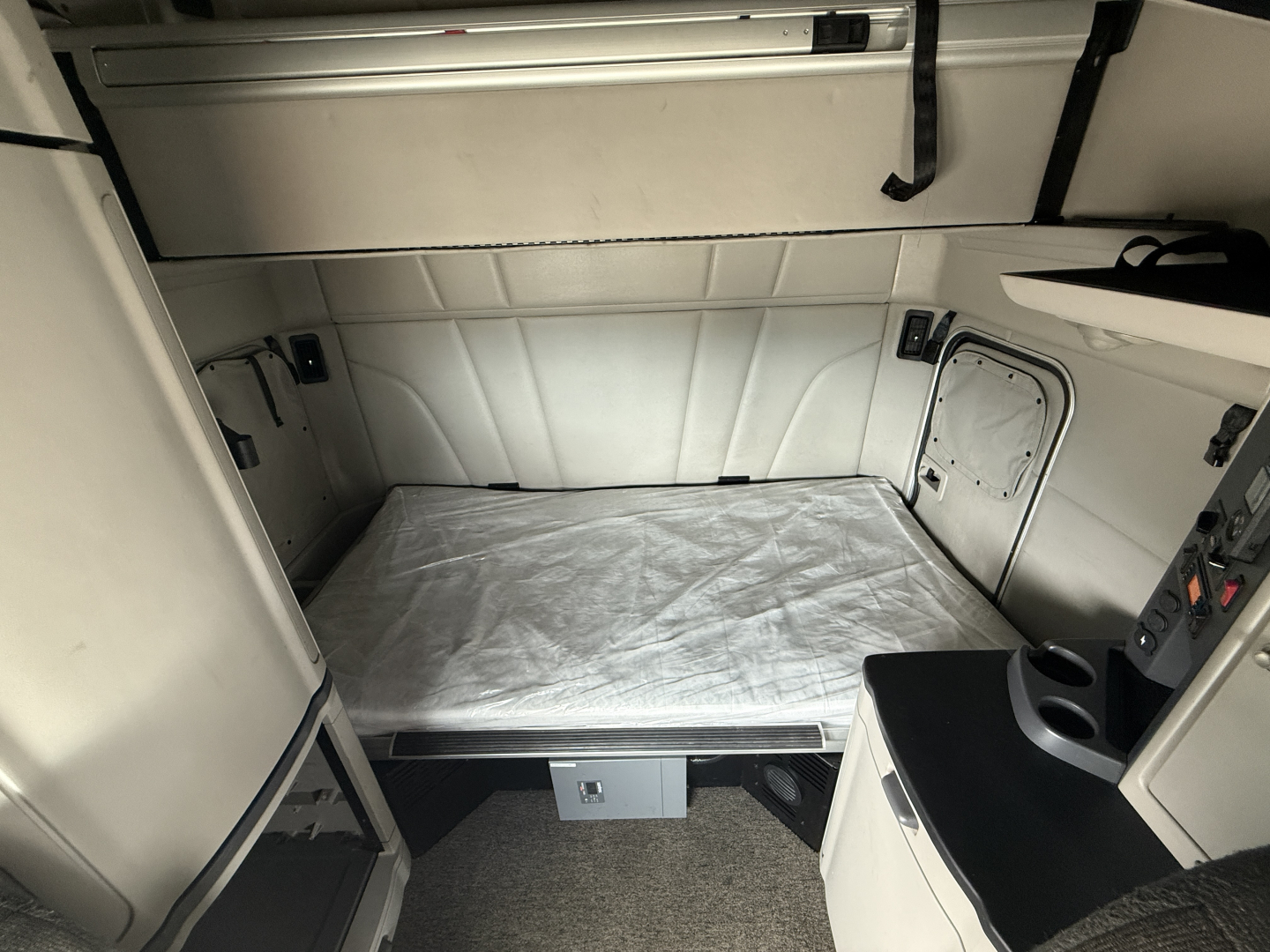Truck sleeper cab interior with a bed platform and storage cabinets in a Peterbilt 579, light gray upholstery and control panel visible.
