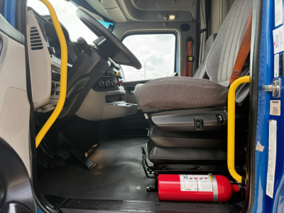 Cab interior of a Peterbilt 579 truck showing driver seat, steering wheel, dashboard, and a red fire extinguisher on the floor near the door.