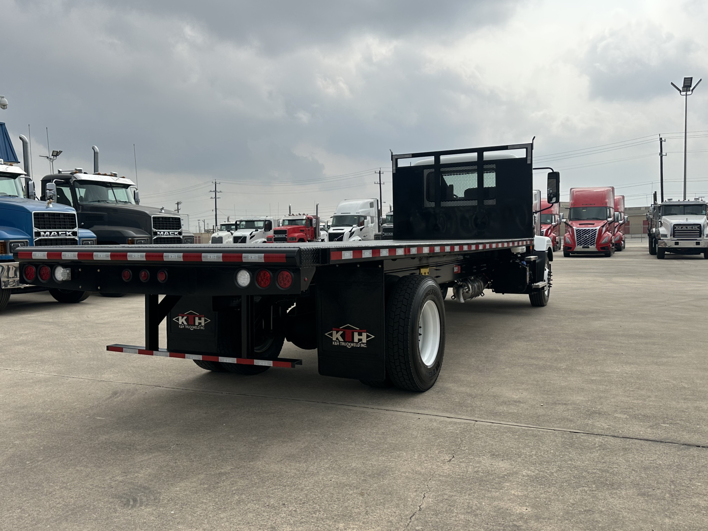 Flatbed truck trailer with empty bed, Mack MD6 not visible, multiple trucks in lot in background.