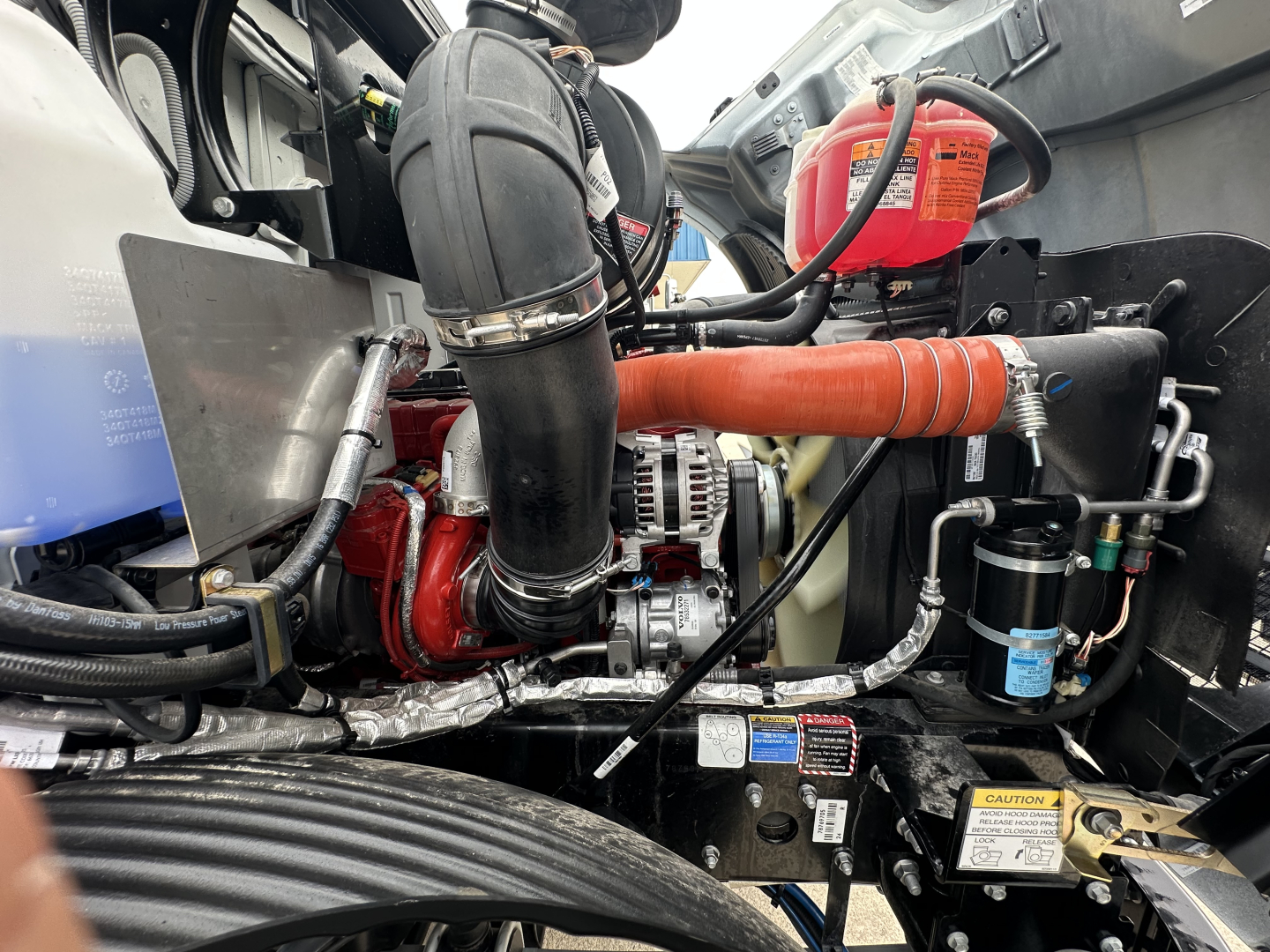 Mack MD6 engine bay view showing turbo intake hose, alternator, orange radiator hose, coolant reservoir, and various mechanical lines in a flatbed truck.