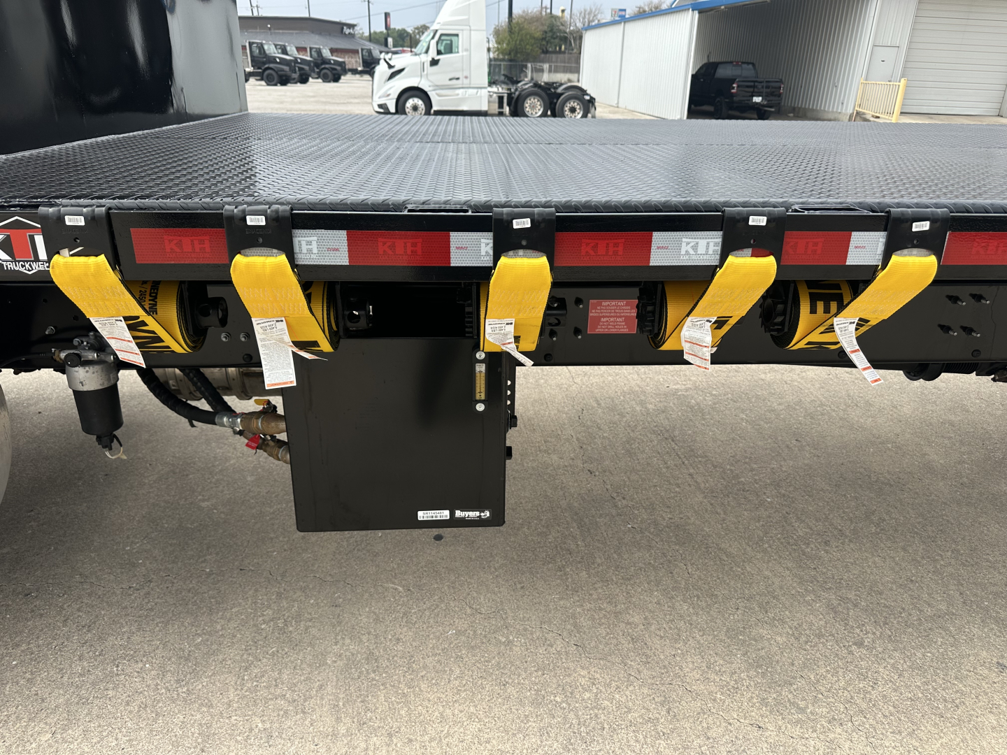 Flatbed trailer rear showing yellow latches and black control box with white tags, truck yard background.