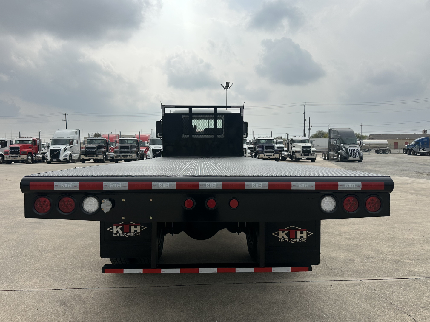 Mack MD6 flatbed truck rear view showing flatbed with dual rear wheels, red/white reflective tape, taillights, mud flaps with KIH logo, and rear bumper.
