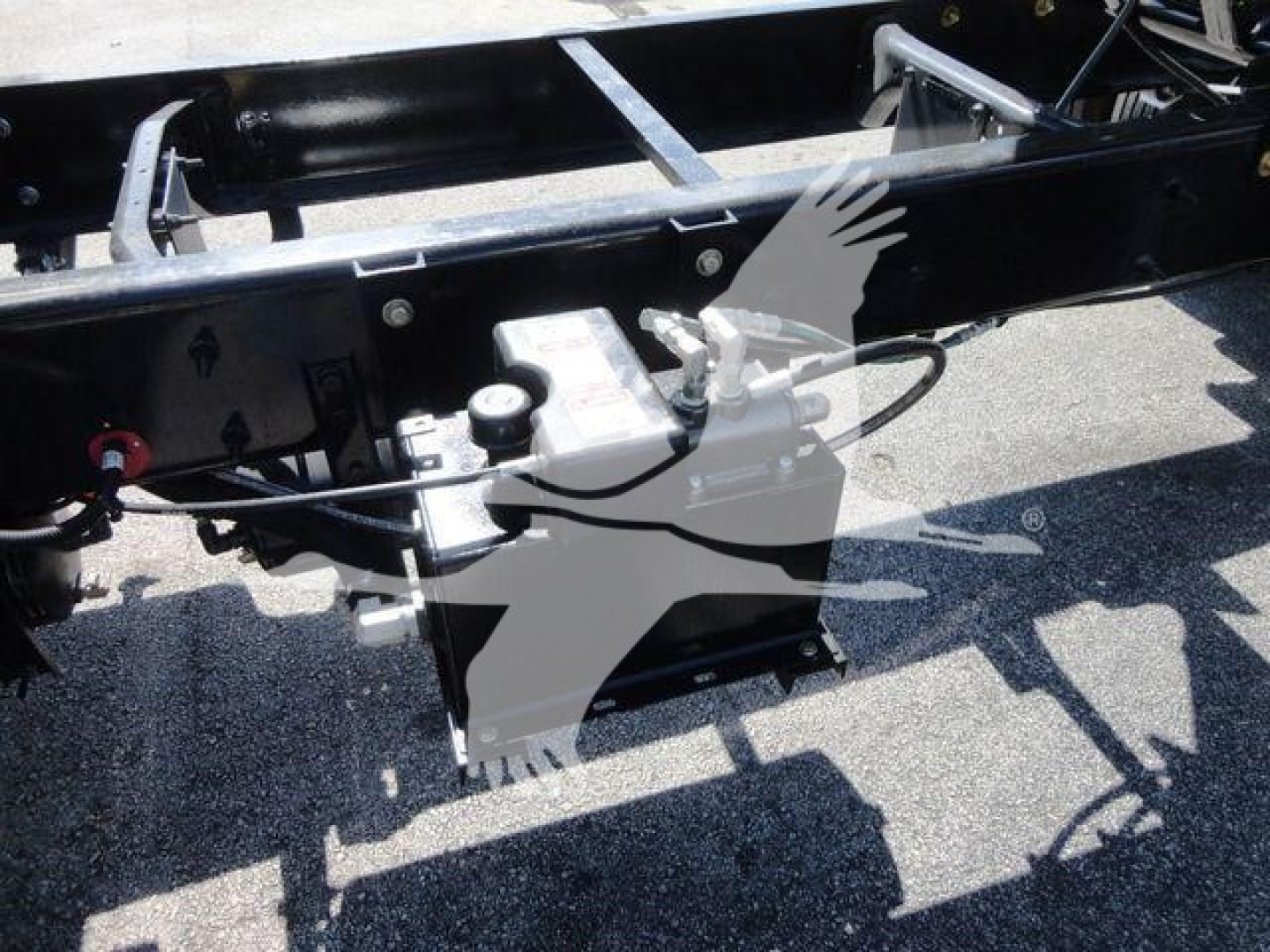 Truck chassis showing hydraulic reservoir and control lines at ground level with frame rails and components.