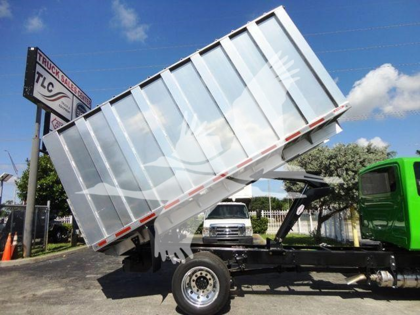 Truck chassis with tilted silver dumping bed in a yard, International MV cab in green visible on right.
