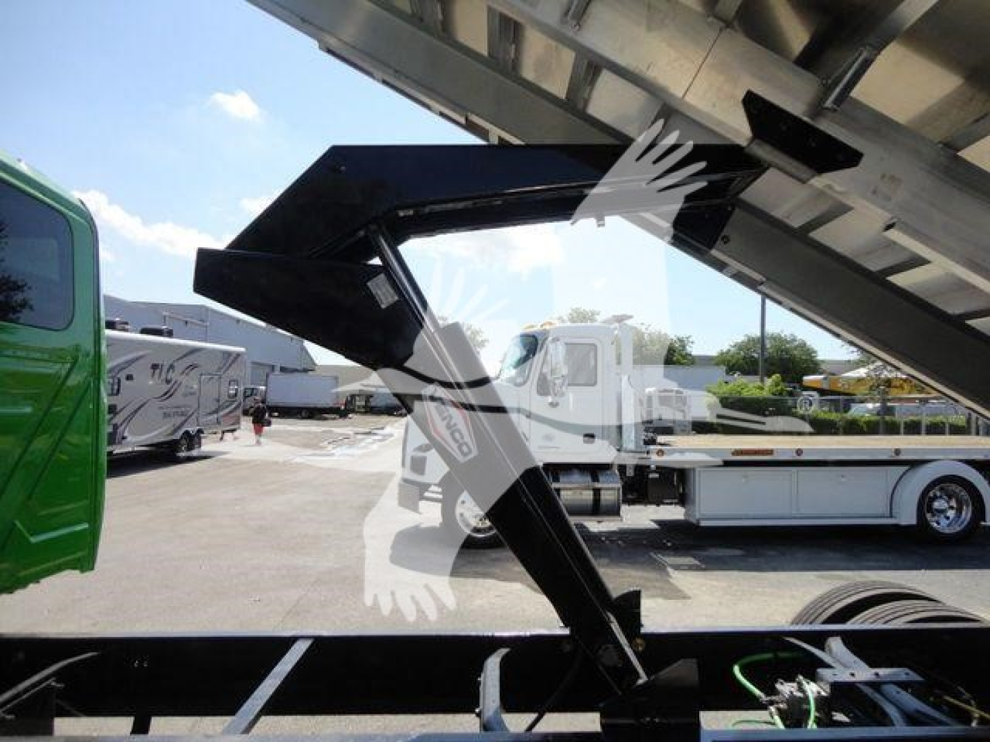 Chipper truck components: hydraulic arm extending from a truck bed, green cab on left, chrome and black metal framework visible, background shows trailers and a clear sky.