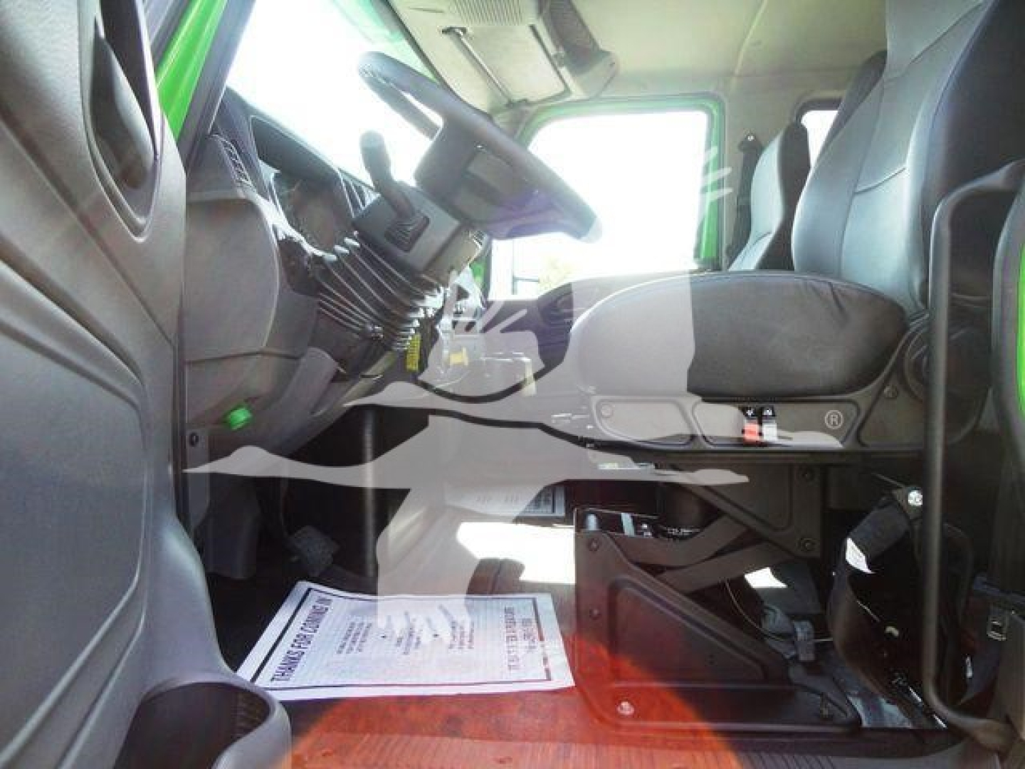 Interior of a 2023 International MV truck cab showing driver seat, steering column, pedals, and dashboard with a document on the floor.