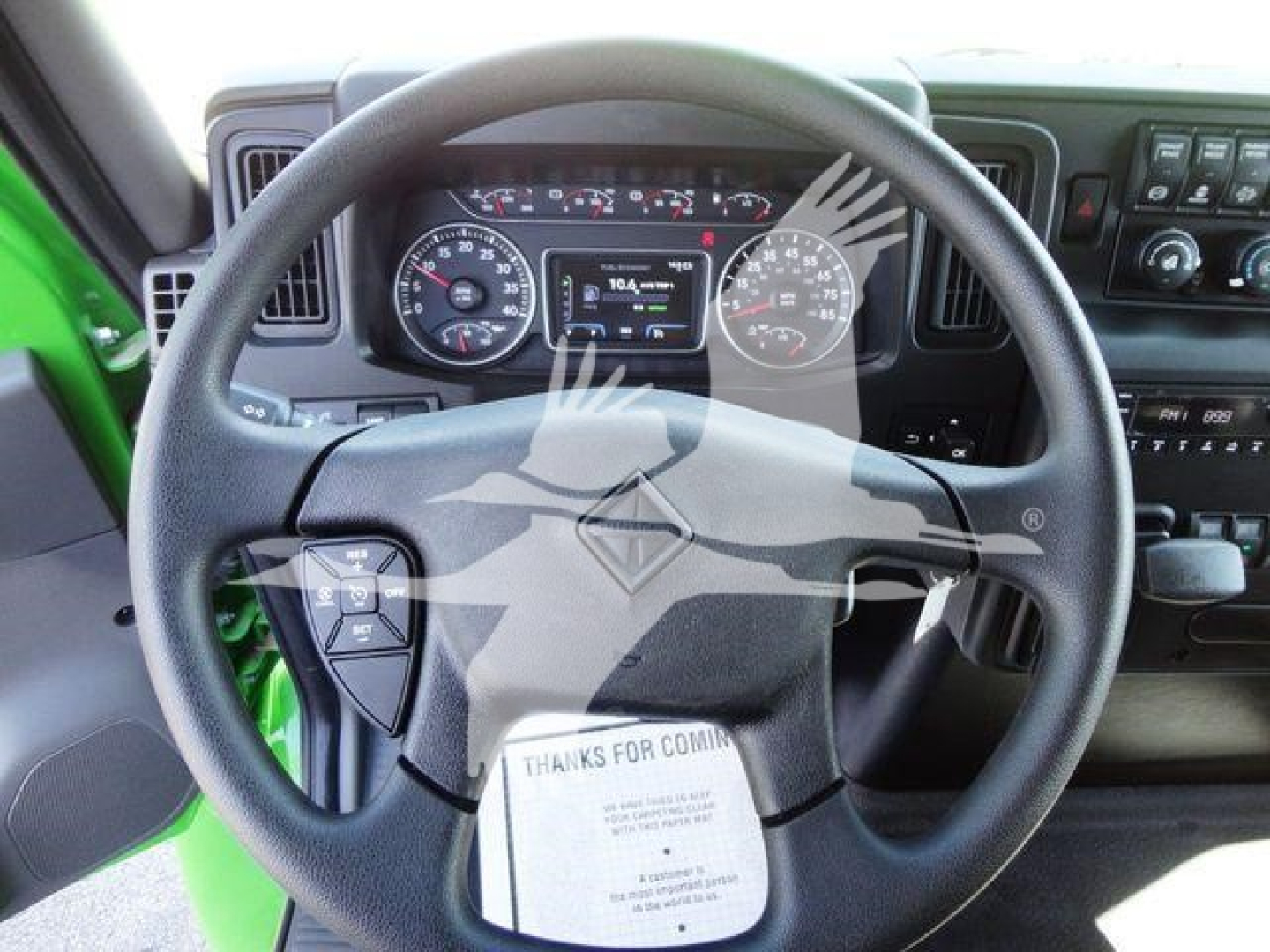 Dashboard and steering wheel of a green 2023 International MV truck showing the instrument cluster and center console. The steering wheel has a prominent diamond logo; a note or card is on the lower part of the wheel.