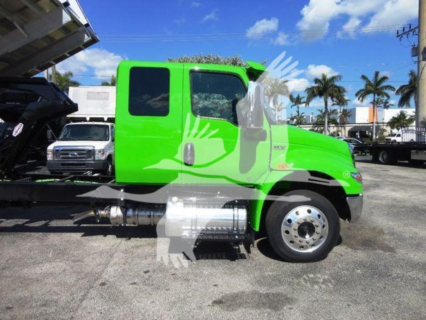Truck cab side profile showing bright green International MV chassis with chrome fuel tanks and wheel, flatbed/chipper equipment visible, dealership lot in background.