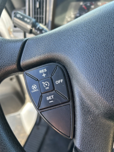 Steering wheel close-up with cruise control buttons visible on left, including RES+/SET-, CANCEL, ON/OFF, and cruise switch cluster.