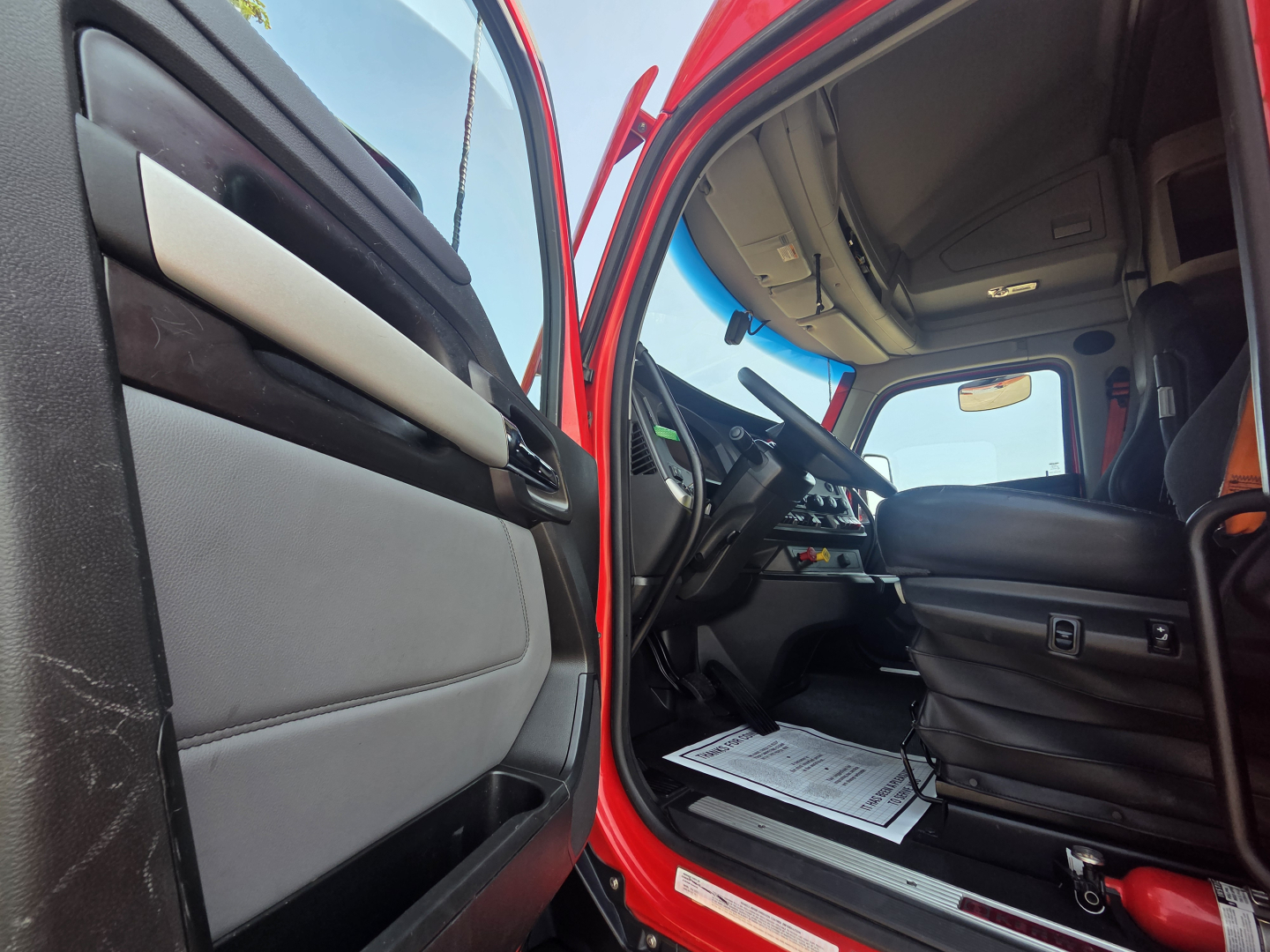 Truck cab interior of a 2024 Kenworth T680 with red exterior, driver seat, steering wheel, and dashboard visible from open driver door.