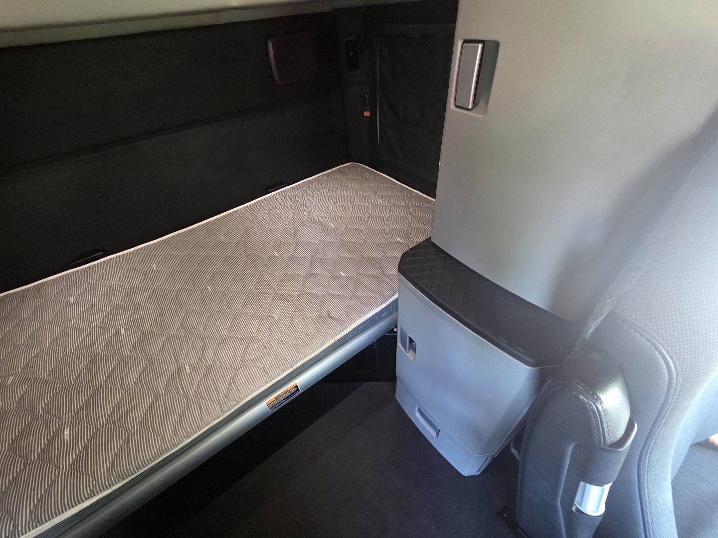 Truck sleeper cab interior showing a bed platform with quilted mattress, side storage cabinet, and cabin wall panel.