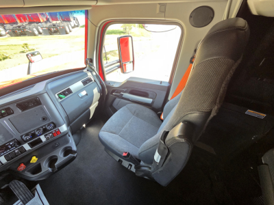 Cab interior of a Kenworth truck with grey seats, dashboard controls, steering wheel, and orange exterior visible through window.