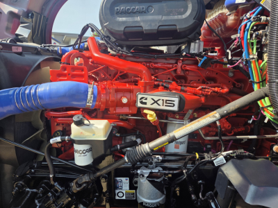 Kenworth T680 engine bay showing  AX15 engine with red intake manifold, blue intercooler hose, Paccar badge, and various fluid reservoirs and wiring harnesses.