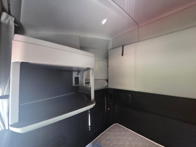 Kenworth T680 sleeper cab interior showing side storage shelf and overhead compartments, with light-colored upholstery and a small window opening.