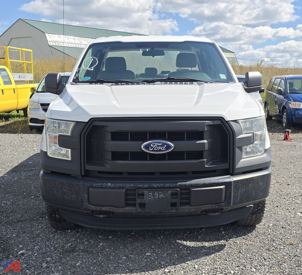 Front view of a white 2015 Ford F-150 pickup truck. Black grille with Ford emblem, large bumper, and headlights; license plate area blank. Weather: clear sky, gravel lot.
