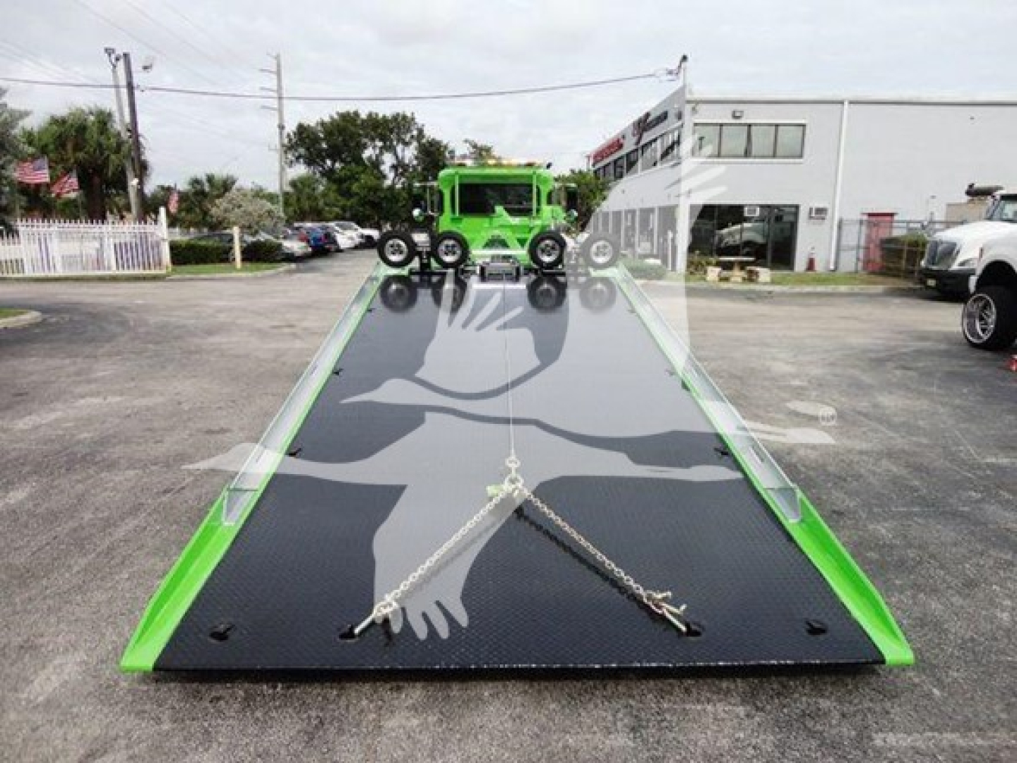 Truck transport trailer bed with green tow truck on top, tilt bed, reflectivity, and chains securing the load. Visible surroundings show dealership lot.