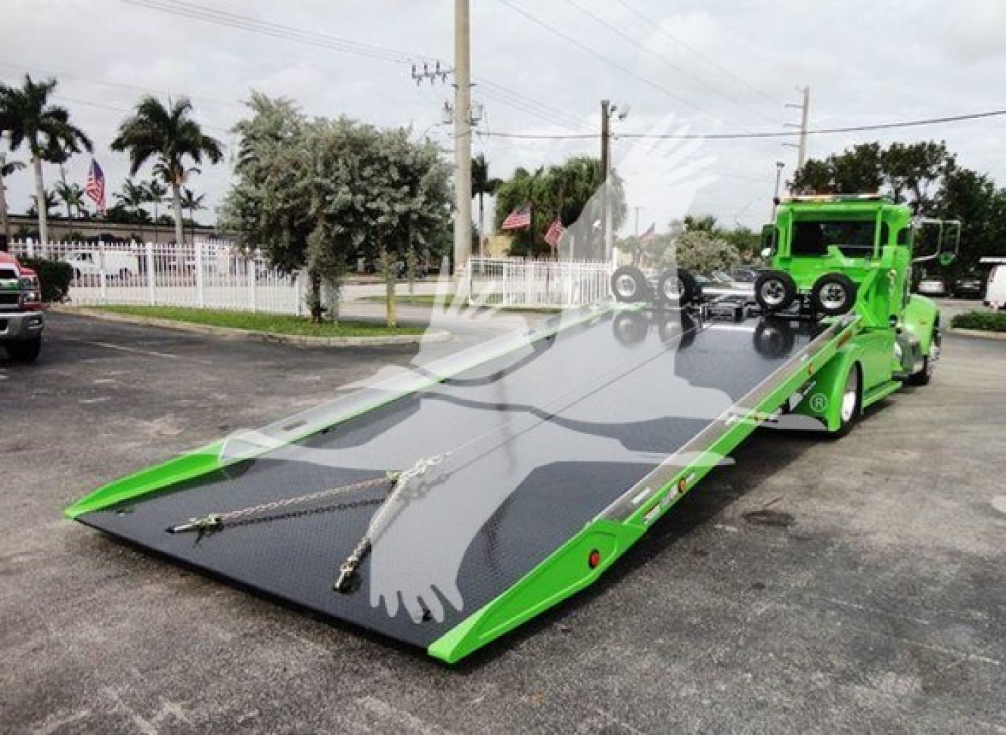 Truck deck showing a bright green rollback tow truck with a long flatbed and winch; license not visible; rear ramp extended.