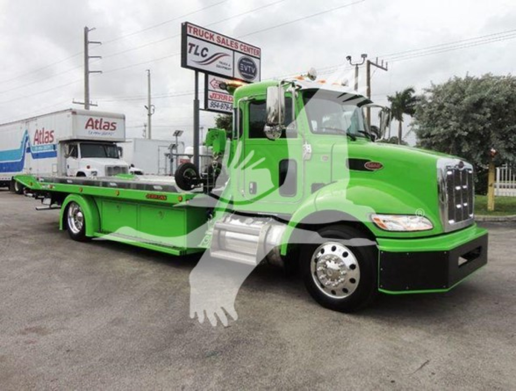 Green Peterbilt 337 rollback wrecker tow truck with flatbed, cab side text not legible, building and trailers in background.