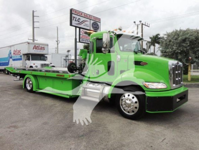 Green Peterbilt 337 rollback wrecker tow truck with flatbed, cab side text not legible, building and trailers in background.