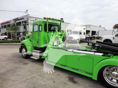 Green Peterbilt 337 rollback tow truck in showroom lot with storage boxes on side, chrome wheels, and white building in background. Visible text on badge on door and side panels.
