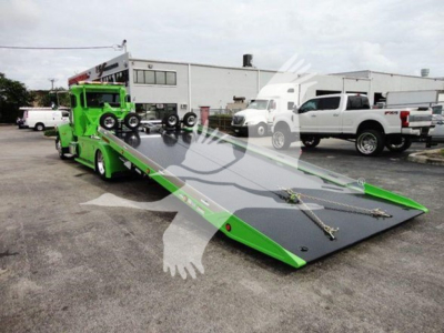 Peterbilt 337 rollback truck with a green tow bed in a dealership lot showing a low-profile flatbed and a white pickup in the background.