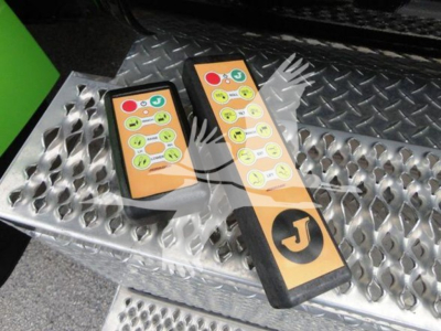 Two remote control panels with multiple colored buttons resting on a diamond-plate step, near a green vehicle body in the background.