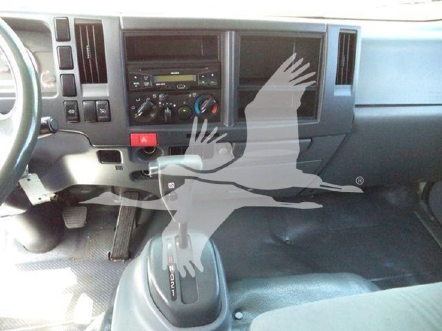 Dashboard area of a 2015 Isuzu NRR truck interior showing steering column, center console with radio, climate controls, and gear shifter. The image includes a translucent watermark of a bird.