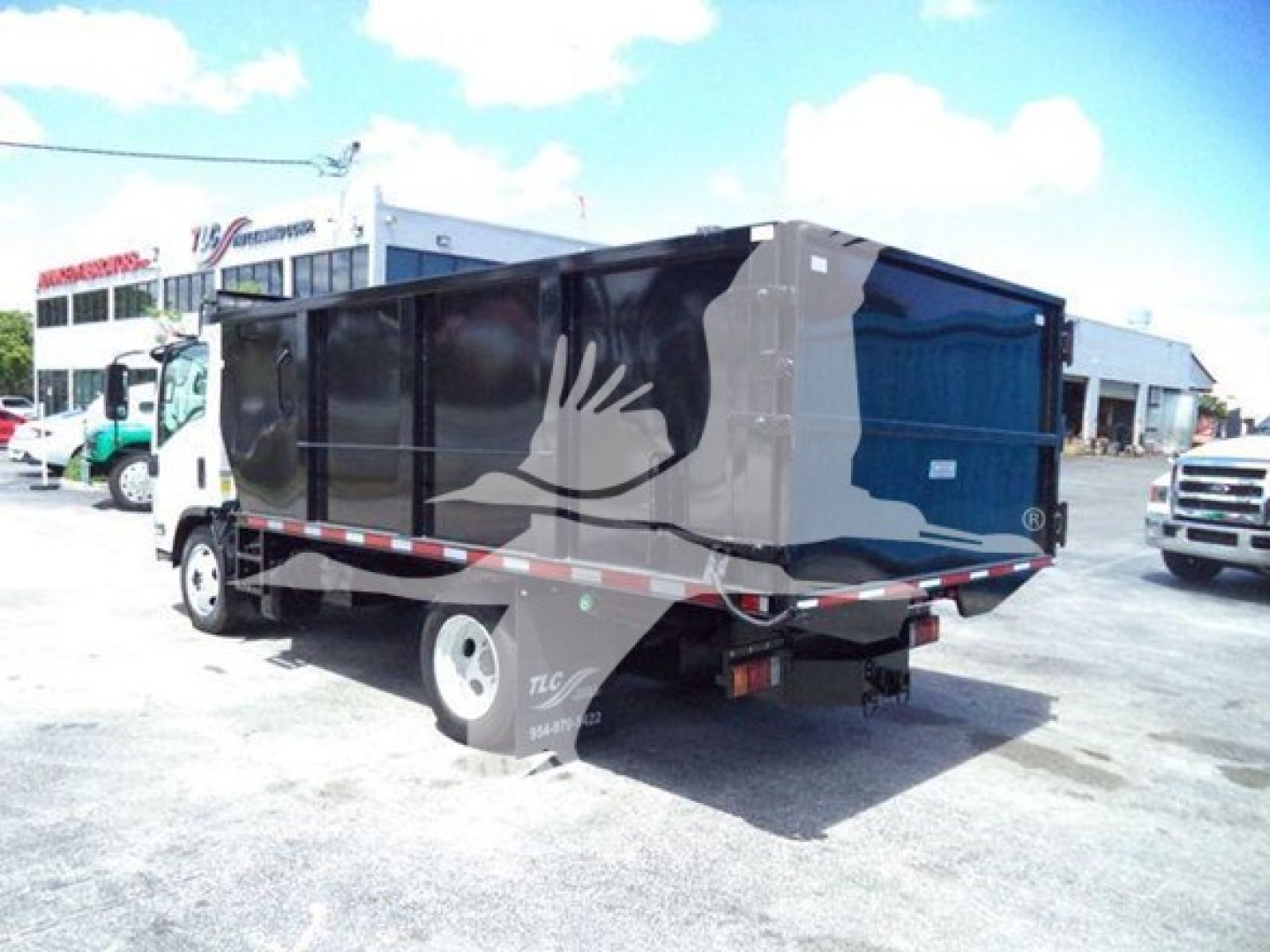 Isuzu NPR/Dump truck rear view showing solid black dump body with logo, taillights, and hitch; warehouse lot in background.