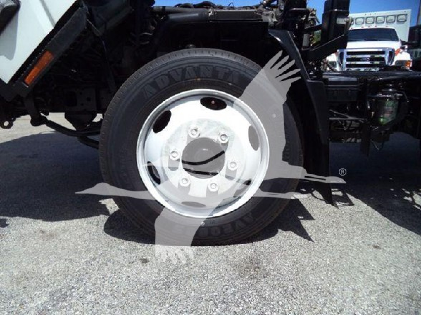 Isuzu NRR truck front wheel and suspension visible; tire shows 'ADVANT' lettering on sidewall; white steel wheel with central hub and lug nuts; bodywork and chassis seen in background.